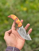 A hand holds the Texan Knives Custom Handmade Damascus Steel Guthook Skinning Knife with a 7.5" blade, camel bone handle, and leather sheath, set against a blurred green background.