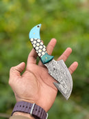 A hand holds the Texan Knives Custom Handmade Damascus Steel Guthook Skinning Knife – Resin Handle 7.5” with colorful blue, cream, and black hexagonal designs. The patterned blade is showcased against a backdrop of blurred greenery.