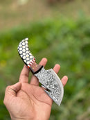 A hand holds the Texan Knives Custom Handmade Damascus Steel Guthook Skinning Knife with a 7.5" patterned blade and a rosewood & resin handle; blurred green foliage is in the background.