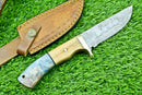 The Texan Knives Handmade Damascus Steel Hunting Knife features a patterned blade, an olive wood & resin handle, and comes with a brown stitched leather sheath with a button closure, displayed on green grass.