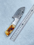 The Texan Knives Damascus Steel Fixed Blade Knife, 7.5" long with a patterned blade and a camel bone & rosewood handle, is shown beside a metal ruler on a light textured surface; includes a leather sheath.