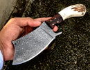 A hand holds the Texan Knives Handmade Damascus Steel Cleaver Knife with an antler horn handle and rosewood bolster. The patterned blade gleams, while a leather sheath is visible in the speckled outdoor background.