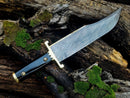 The Texan Knives 13-Inch Damascus Steel Cowboy Bowie Knife, featuring a buffalo horn handle and leather sheath, rests on weathered wood with green moss. Its intricate wavy blade pattern is clearly visible—a perfect gift for knife enthusiasts.