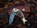 The Texan Knives Custom Handmade Fish Style Damascus Steel Folding Knife features a sheep horn and brass handle, designed to resemble a fish, and comes with a leather sheath displayed on dark, textured wood.