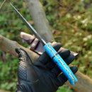 A gloved hand holds the Texan Knives Custom Handmade Damascus Steel Hunting Knife – 10-Inch Blade with resin handle; outdoors, greenery and the included leather sheath are visible in the background.