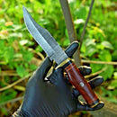 A hand in a black glove holds the Texan Knives Damascus Steel Hunting Knife—10" blade with brass guard and resin pakkawood handle—against blurred green foliage. Leather sheath included.