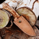 The Texan Knives Exquisite 10-Inch Damascus Steel Hunting Knife, featuring a rosewood handle and leather sheath, is displayed on stacked logs—an ideal gift for outdoorsmen.
