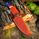 The Texan Knives Custom Handmade Damascus Skinning Knife features a vibrant American flag handle and rests in a brown leather sheath on a cut log outdoors.