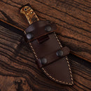 The Texan Knives Custom Handmade Damascus Steel Skinning Knife features a 4.5" blade and a sheep horn handle, shown partially inside a dark brown leather sheath with white stitching and black snap buttons, resting on a wooden surface.