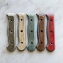 Five Messermeister Custom Driftwood Mod Kit Handles (Small) in gray, cream, blue, brown, and red—each with three holes—are arranged vertically on a white marble surface for your custom knife upgrade.