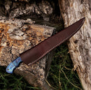 The Texan Knives Handmade 8" Rasp Steel Sheep Horn Hunting Knife with belt sheath lies outdoors on rough-cut logs and grass.