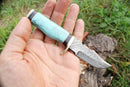 A hand holds the Texan Knives Custom Handmade Best Survival Knife featuring a patterned Damascus steel blade and a light blue marbled handle, with grass and wood in the outdoor background. Brand: Texan Knives.