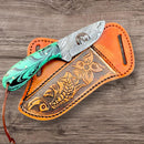 The Texan Knives Custom Handmade Bull Cutter Knife with Sheath features a green and black handle and rests on a brown leather sheath engraved with floral and fish designs, displayed on a wooden surface.