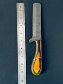The Texan Knives Bull Cutter Knife, a handmade Damascus steel blade with a black-and-orange pakkawood handle, is shown beside a metal ruler on a dark surface. This custom knife measures approximately 10 inches in length.