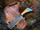 The Texan Knives Custom Handmade Bull Cutter, featuring a Damascus steel blade and a wooden handle with blue and metal accents, rests on its brown leather sheath with decorative stitching on wet rocks.