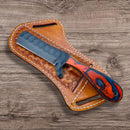 The Texan Knives Custom Handmade Bull Cutters feature an 8-inch J2 steel blade and a patterned orange and black wooden handle, shown resting on a brown leather sheath atop a wooden surface.