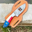 The Texan Knives Patriotic Custom Handmade Cowboy Knife, featuring a USA flag handle and 8" Damascus blade, rests on its tan leather sheath. The branded knife displays a decorative pattern and cut-out near the hilt by grass and water.