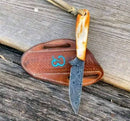 The Texan Knives Custom Handmade Damascus Belt Knife with a rust-colored camel bone handle and patterned blade rests on a brown leather sheath with a blue swirl, both displayed on weathered wooden planks.