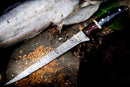 The Texan Knives Handmade Damascus Fillet Knife 13” with a buffalo horn, rosewood, and brass handle rests on wood beside a partially filleted fish and green leaves, its leather sheath nearby amidst wood shavings.
