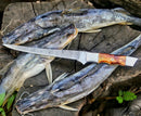 Three raw fish with whisker-like fins rest on weathered wood, a Texan Knives Custom Handmade Damascus Filleting Knife – 13" Camel Bone Handle with Leather Sheath placed across them. A small green fern decorates the lower right corner.