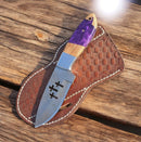 The Texan Knives Custom Handmade Damascus Skinner Knife features a patterned blade with three cross-shaped cutouts and an olive wood handle with purple resin, resting on a woven leather sheath for outdoor and hunting use.
