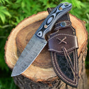 The Texan Knives Custom Handmade Damascus Skinning Knife, featuring a 4.5” patterned blade and black-and-gray handle, rests beside its brown leather sheath on a tree stump with green foliage in the background.