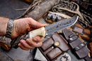 A hand holds the Texan Knives Handmade Damascus Skinning Knife with a polished camel bone and sheep horn handle. Behind it, leather pouches, shotgun shells, rope, and outdoor gear create a rugged, rustic scene.