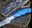 A Custom Handmade 13-Inch Damascus Steel Fillet Knife Blank with a resin handle from Texan Knives rests on a tree stump next to a catfish head, its patterned blade and premium leather sheath visible.