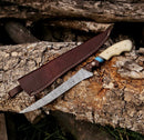 The Texan Knives Fishing Fillet & Boning Knife features a 13" Damascus steel blade with a camel bone and rosewood handle, resting on a log beside its brown leather sheath.