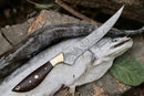 A 13" Damascus Fillet Knife by Texan Knives, featuring a rosewood handle and leather sheath, rests atop a large silver fish on a tree stump with a green leaf nearby.