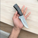 A hand holds the Texan Knives Custom Handmade Fixed Blade Cowboy Knife with an authentic sheep horn handle and shiny blade, over a light wood surface.
