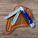 The Texan Knives Folding Bull Cutter Knife – Custom Handmade Damascus Steel, featuring two open blades and a blue handle, is shown on a brown tooled leather sheath atop a wooden surface.
