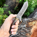 A hand holds the Texan Knives Whiskey Knives Out, featuring a 4-inch Damascus steel blade and sheep horn handle, displayed outdoors near a tree stump with green foliage, beside its finely crafted leather sheath.