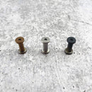 Three Messermeister Custom Mod Kit Handle Hardware screws in bronze, silver, and black—perfect for upgrading your knife handle—are arranged in a row on a textured, light gray surface.