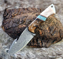 The Texan Knives Premium Damascus Guthook Skinning Knife features a patterned steel blade and rosewood & bone handle, displayed on textured wood and fur—an essential 10” hunting knife.