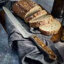A Messermeister bread knife with a Custom Russet Mod Kit Handle - Large rests on a gray cloth beside slices of multigrain bread on a wooden cutting board.