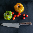 A Messermeister Custom Russet Mod Kit Handle - Small chef’s knife by Messermeister rests on a dark surface beside a green tomato, a yellow tomato, and small red cherry tomatoes.