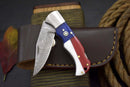 The Texan Knives Handcrafted Damascus Steel Pocket Knife features a Texas flag handle with a gold star and comes with a brown leather sheath. A coiled rope, striped surface, and red leaf add to the scene’s rustic charm.