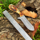 The Whisky Bent Knives – Custom Handmade Damascus Steel Blade by Texan Knives features a patterned blade and carved wooden handle, shown on tree bark beside a dual-scale metal ruler and accented with green leaves.