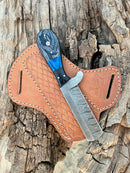 The Texan Knives Custom Handmade Bull Cutter Knife – Damascus Steel Holiday Edition, featuring a patterned blade and black-blue handle, rests on a stitched brown leather sheath atop weathered wood—a stunning limited piece by Texan Knives.