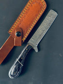 The Texan Knives Handmade Damascus Steel Calf Cutter Knife features a 5" straight-edged blade and a pakkawood handle, resting next to its brown leather sheath with yellow stitching.