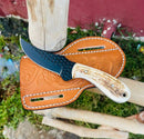 The Texan Knives Cowboy Bull Cutter Knife features a handmade J2 steel blade and stag horn handle, resting on a tooled brown leather sheath outdoors on wooden poles, with grass and a red wall in the background.