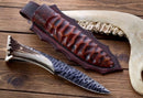 The Texan Knives Custom Forged 1095 High Carbon Steel Hunting Knife with a textured blade, antler horn handle, and handcrafted leather sheath rests on wood; an animal jawbone appears in the background.