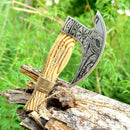 The Texan Knives Custom Hand Engraved Ax Pizza Cutter with a J2 steel blade features a carved wooden handle and is displayed embedded in weathered wood outdoors, surrounded by lush green foliage.