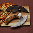 The Texan Knives Axe Pizza Cutter—custom hand-engraved with a J2 steel blade and wooden handle—rests on a cutting board with a partially sliced pizza, leather sheath nearby, all set against a reddish-brown background.