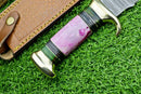 A close-up of the Texan Knives Damascus Steel "The Bowie" Knife—16" with a brass guard and marbled resin pakkawood handle, plus leather sheath—all displayed on green artificial grass.