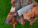 The Texan Knives Damascus Steel Hunting Knife – 10" with Rosewood Handle and Leather Sheath is displayed on tree bark amid green grass, highlighting its patterned blade and polished finish.