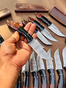 A hand holds the Texan Knives Custom Handmade 4.5-Inch Damascus Steel Skinning Knife with a resin and rosewood handle, while similar Texan Knives and brown leather sheaths are displayed on the surface in the background.