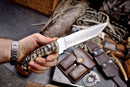 A hand holds the Texan Knives Handmade J2 Steel Skinner Knife with a sheep horn handle and 4.5-inch blade; leather sheaths, a watch, a box, and various outdoor survival gear are seen in the background.