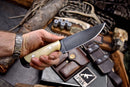 A person holds a Texan Knives Custom Handmade J2 Steel Skinning Knife with a 4.5" camel bone handle; outdoor gear like rope, shotgun shells, and pouches are laid out on a wooden surface in the background.